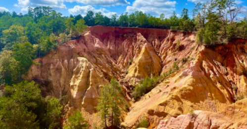 Colorful, layered rock formations surrounded by green trees under a blue sky with fluffy clouds.
