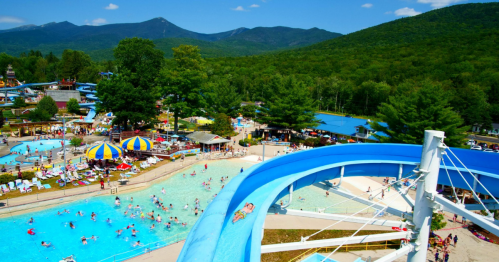 Aerial view of a vibrant water park with slides, pools, and visitors enjoying a sunny day surrounded by mountains.