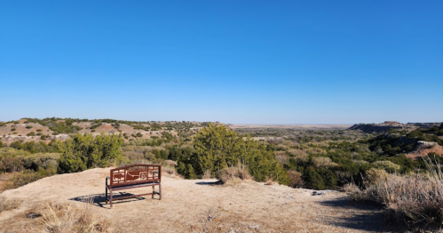 A metal bench overlooks a vast landscape of rolling hills and trees under a clear blue sky.