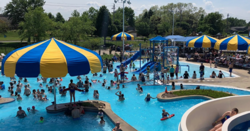 A busy water park with colorful umbrellas, slides, and many people enjoying the pool on a sunny day.