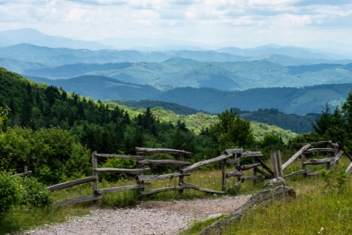 A wooden fence along a path overlooks lush green mountains and rolling hills under a cloudy sky.
