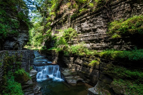 A serene landscape featuring a narrow gorge with lush greenery and a gentle waterfall flowing through rocky cliffs.