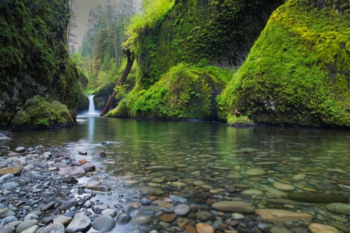 A serene river flows through lush green cliffs, with a small waterfall in the background and smooth stones lining the shore.