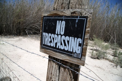 A weathered wooden post with a bold "No Trespassing" sign, surrounded by dry grass and a barbed wire fence.