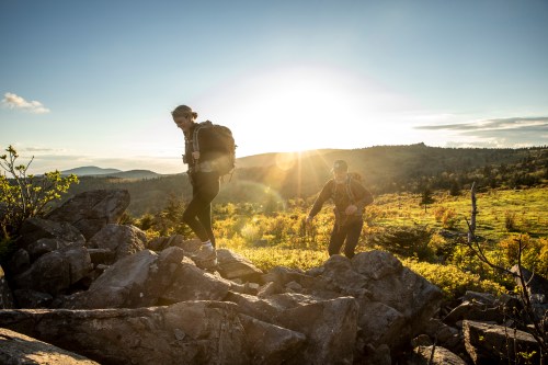 Two hikers navigate rocky terrain at sunset, with a vibrant landscape and mountains in the background.