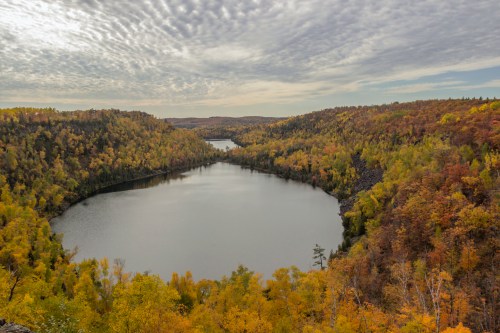 A serene lake surrounded by vibrant autumn foliage and rolling hills under a cloudy sky.