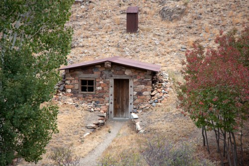 A small stone cabin with a wooden door, surrounded by dry grass and trees, set against a rocky hillside.
