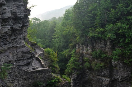 A winding path through lush green trees and rocky cliffs in a serene, misty landscape.
