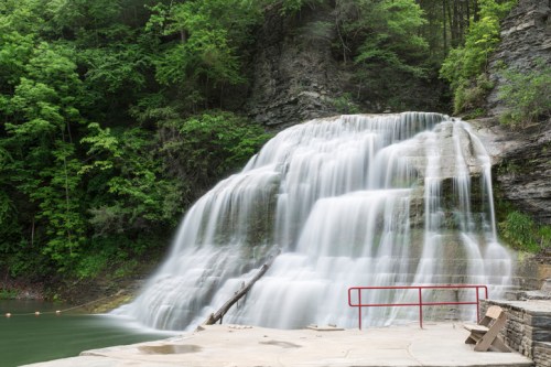 A serene waterfall cascades over rocks, surrounded by lush green trees, with a red railing in the foreground.
