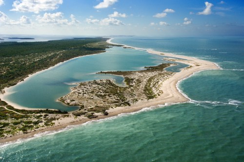 Aerial view of a coastal landscape featuring sandy shores, lush greenery, and calm blue waters under a partly cloudy sky.
