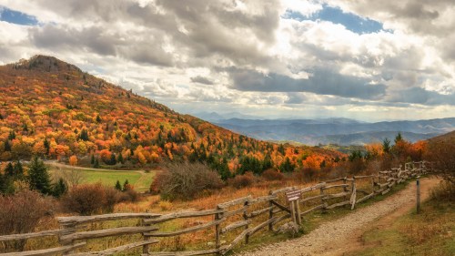 A scenic view of a mountain landscape in autumn, featuring vibrant orange and yellow foliage under a cloudy sky.