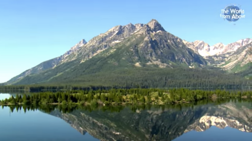 A serene mountain landscape reflected in calm water, surrounded by lush greenery and distant snow-capped peaks.