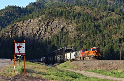 A train passes by a sign for a fishing access point, with mountains and trees in the background.