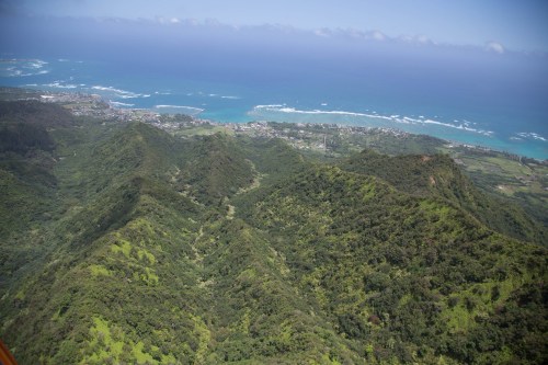 Aerial view of lush green mountains leading to a coastline with blue ocean waves and a distant town.