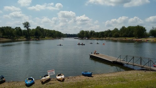 A serene river scene with kayakers paddling, a dock, and lush greenery under a partly cloudy sky.