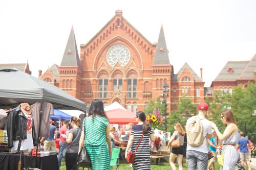 People gather at an outdoor market with tents, food stalls, and a historic red brick building in the background.