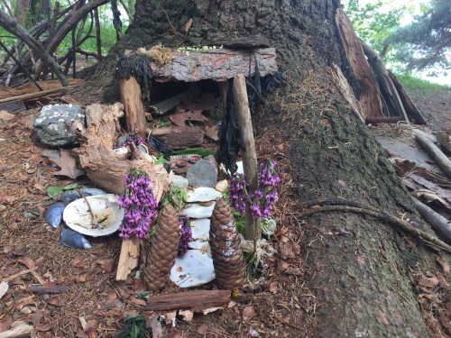 A small, whimsical structure made of natural materials, featuring pinecones and flowers, nestled at the base of a tree.