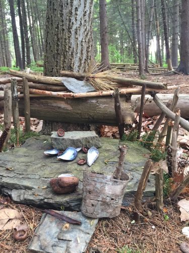 A small, rustic shelter made of sticks and stones, nestled against a tree in a forest setting.