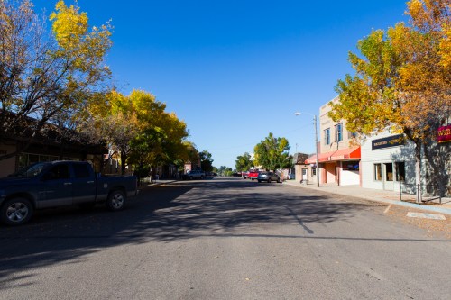 A quiet street lined with trees displaying autumn colors, featuring shops and parked cars under a clear blue sky.