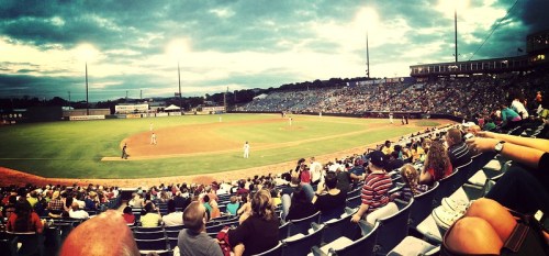 A panoramic view of a crowded baseball stadium under cloudy skies, with players on the field and fans in the stands.