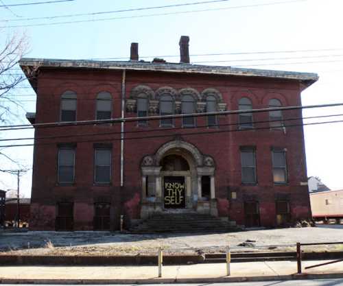 Abandoned red brick building with a large archway and a sign reading "KNOW THY SELF" hanging from the entrance.