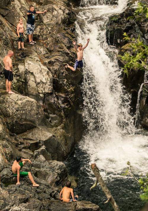A group of people near a waterfall, with one boy jumping off a rock into the water below.
