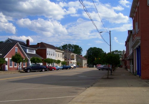 A quiet street lined with trees, featuring parked cars and buildings under a partly cloudy sky.