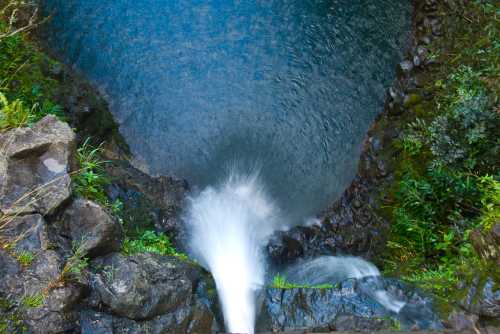 Aerial view of a waterfall cascading into a deep blue pool surrounded by lush greenery and rocky terrain.