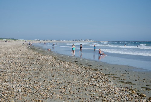A sunny beach scene with people walking along the shore and playing in the water, surrounded by pebbles and waves.