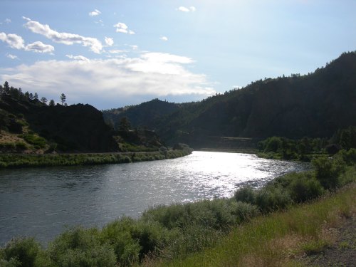 A serene river flows through a lush valley, surrounded by mountains and a bright blue sky with scattered clouds.