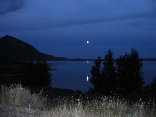 A serene night scene with a full moon reflecting on a calm lake, surrounded by mountains and trees.
