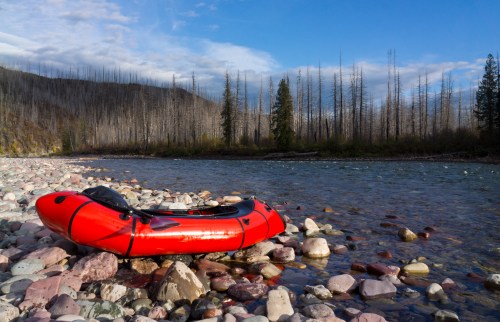 A red inflatable kayak rests on a rocky riverbank, surrounded by trees and a clear blue sky.