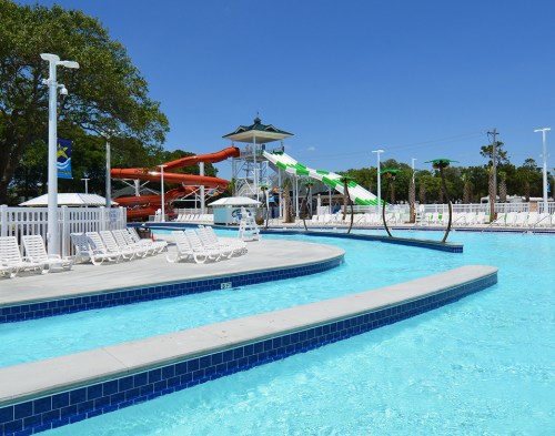 A bright outdoor pool area featuring water slides, lounge chairs, and clear blue water under a sunny sky.
