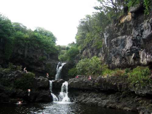 A serene waterfall cascades into a rocky pool, surrounded by lush greenery and people enjoying the natural setting.