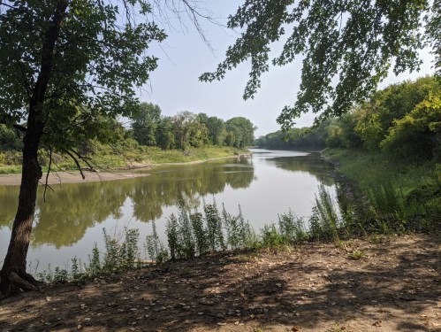 A calm river surrounded by lush greenery and trees, reflecting the clear sky on a sunny day.