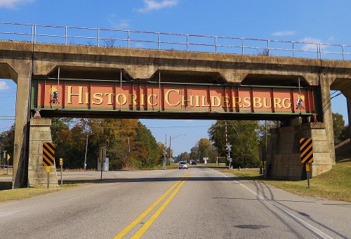Sign reading "Historic Childersburg" on a bridge over a road, surrounded by trees and clear blue sky.