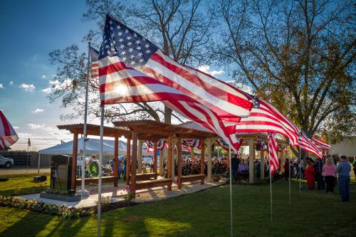 American flags wave in the breeze at an outdoor event with a gathering under a wooden pavilion and trees in the background.
