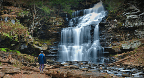 A person stands near a cascading waterfall surrounded by rocky terrain and lush greenery.