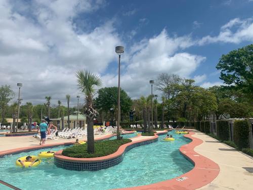 A sunny pool area with a winding lazy river, palm trees, and lounge chairs under a partly cloudy sky.