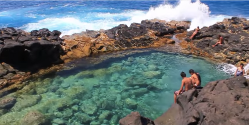 A natural rock pool by the ocean, with people sitting on the rocks and waves crashing nearby.