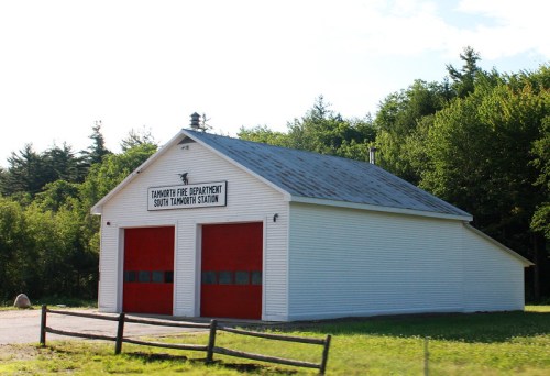 A white fire station building with red doors, surrounded by trees and a wooden fence. Sign reads "Tamworth Fire Department."