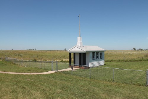 A small white chapel with a cross on top, surrounded by a fence and open grassy fields under a clear blue sky.