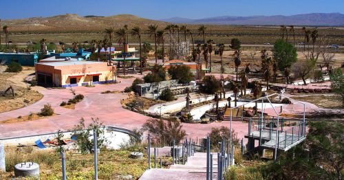 Abandoned amusement park with colorful buildings, overgrown palm trees, and empty pathways under a clear blue sky.
