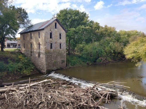 Abandoned stone building beside a river, with trees and a small waterfall in the foreground under a partly cloudy sky.