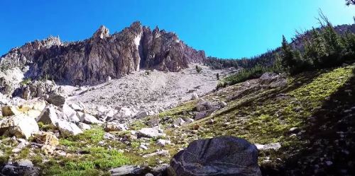 A rocky mountain landscape with steep cliffs, scattered boulders, and green vegetation under a clear blue sky.