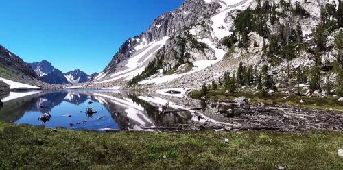 A serene mountain landscape with a clear lake reflecting snow-capped peaks and lush greenery under a blue sky.
