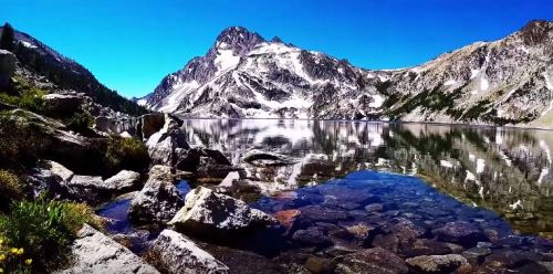 A serene mountain lake reflecting snow-capped peaks under a clear blue sky, surrounded by rocky shores and greenery.
