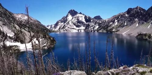 A serene mountain lake surrounded by rugged peaks and snow, under a clear blue sky.