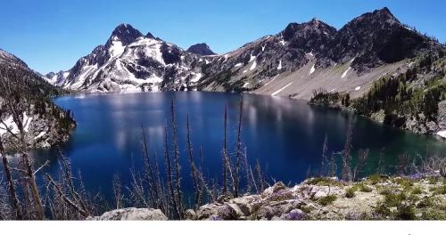 A serene mountain lake surrounded by rugged peaks and clear blue skies, with lush greenery in the foreground.