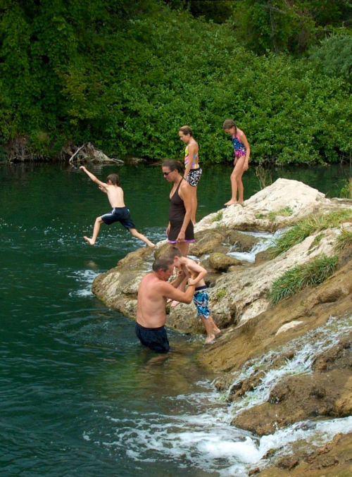 A group of children and adults enjoying a day by the water, with one child jumping in and others playing on the rocks.